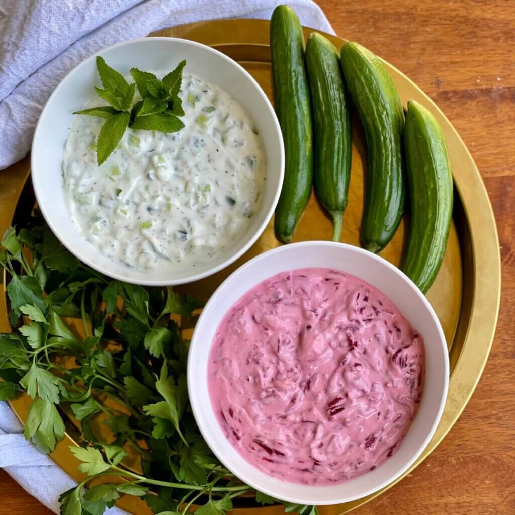 Bowls of Maast o Khiar and beet yogurt dip surrounded by fresh herbs and cucumbers